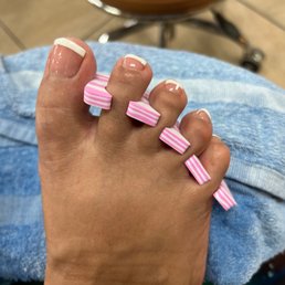 Emma's attention to detail. French manicure on toes. She brings your feet back to life. - The image displays a person's foot with a French pedicure, characterized by white tips on the toenails. The foot is resting on a surface, with a background that includes a chair and tiled flooring. Pink and white striped toe separators are placed between the toes to keep them apart during or after nail treatment. The person is wearing blue denim jeans, and the toenails are well-groomed with a shiny finish.