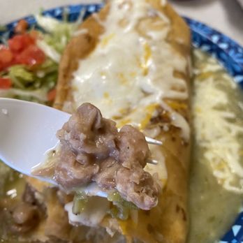 Papa Nachos - Simply fantastic bean and cheese burrito, chimichanga style with green chile sauce - A large burrito covered with melted cheese and sour cream is presented on a blue patterned plate. A spoon holds a portion of refried beans and chopped vegetables. In the background, there are small containers of salsa and sauce, along with a side salad of lettuce and diced tomatoes. The dish appears to be part of Mexican or New Mexican cuisine, served in a casual dining setting. - Mexican Near Me - Albuquerque, New Mexico