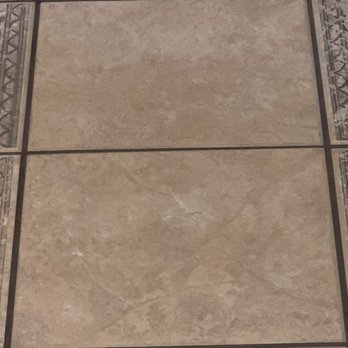Stanley Steemer - Hallway - AFTER - The image shows a section of tiled flooring consisting of large beige ceramic tiles with a textured surface. The tiles are separated by dark grout lines. Decorative border tiles with intricate patterns are visible along the edges of the flooring, framing the central beige tiles. - Carpet Cleaning Near Me - Holbrook, Massachusetts