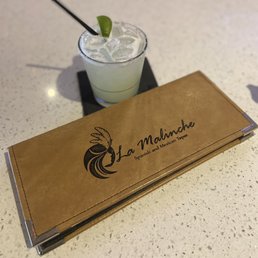 Happy hour Margarita - A margarita cocktail with ice and a lime wedge is placed on a black coaster on a speckled white table. Next to the drink is a closed, brown leather menu with the La Malinche logo and text indicating Spanish and Mexican tapas. The setting suggests a dining environment at La Malinche restaurant.