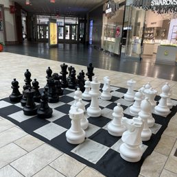 A large outdoor chess set with oversized black and white chess pieces is arranged on a black and white checkered chessboard mat in a shopping mall. The mall features tiled flooring, storefronts such as Swarovski, and an entrance to Paramus Park dental. The chess pieces are made of a sturdy material, and the setup is in an open area near the mall's entrance and retail shops.