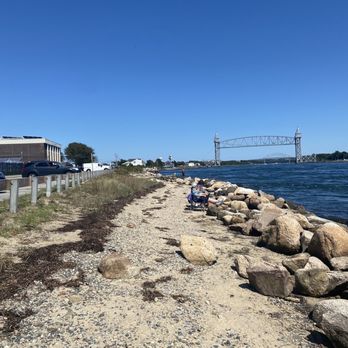 Mass Maritime Academy - A gravel and rocky shoreline extends along a body of water, with several people seated on chairs, facing the water. The scene includes a parking area with cars on the left, a grassy strip, and a bridge structure in the background. The sky is clear and blue, indicating a sunny day. - Colleges & Universities Near Me - Buzzards Bay, Massachusetts