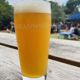 Hazy Ipa - A tall, clear glass of hazy IPA beer with a thick foam head is positioned on a weathered wooden table outdoors. The glass has the 'Meanwhile Brewing' logo printed on it. In the background, there are trees, a blue canopy, and groups of people sitting at picnic tables, indicating an outdoor gathering at a brewery. The sky is partly cloudy.