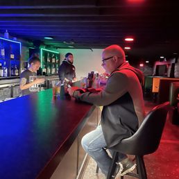 Dani and some construction sales  dude - A man with glasses and a jacket is seated at a bar counter, looking at his phone, with two bartenders behind the bar in a nightclub setting.