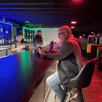 Night Train - Dani and some construction sales  dude - A man with glasses and a jacket is seated at a bar counter, looking at his phone, with two bartenders behind the bar in a nightclub setting. - Cocktail Bars Near Me - Fort Wayne, Indiana