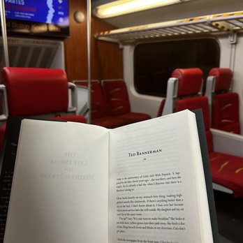 Beastly Books - Catriona Ward - The image shows a person holding an open book titled 'Ted Bannerman' inside a train carriage. The train has red upholstered seats, some of which are empty, and a window revealing a dark night outside. Overhead lighting illuminates the interior, and a digital screen is visible in the background displaying text in Spanish. The person's hand is visible, holding the book, and the scene suggests a quiet moment of reading during a train journey. - Bookstores Near Me - Santa Fe, New Mexico