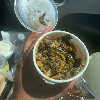 Ray Ray's Hog Pit - Greens - A person with dark skin holds two cups of food inside a vehicle. One cup contains cooked greens, and the other appears to have a beverage with ice and small food particles. The background shows a water bottle, a container of coleslaw, and some food packaging, indicating a meal or takeout setting. - Barbeque Near Me - Westerville, Ohio