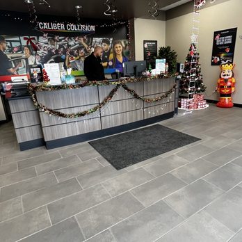 Caliber Collision - Their front desk - The image shows the reception desk of Caliber Collision, decorated for the holiday season with Christmas garlands, ornaments, and festive decorations. A small Christmas tree and a nutcracker figure are positioned near the wall, and hanging decorations include spiraled ribbons and holiday-themed items. The background features a large photo mural of young people and a sign reading 'Caliber Collision.' The floor is tiled, and the overall setting is a modern, welcoming reception area with holiday cheer. - Body Shops Near Me - Rio Rancho, New Mexico
