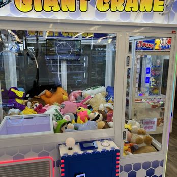 Claw Party Arcade - A claw machine at Giant Crane arcade in Glendale, AZ, contains numerous plush toys of different characters and animals. The machine has a glass enclosure with a control panel at the front, featuring a joystick and buttons. The arcade environment includes other gaming machines and bright signage, with the Giant Crane sign prominently displayed at the top. The plush toys inside include recognizable characters and generic stuffed animals, arranged in a pile for players to attempt to grab. - Arcades Near Me - Glendale, Arizona