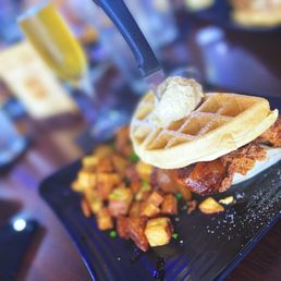 Chicken and Waffles - A close-up of a chicken and waffle dish featuring a crispy fried chicken piece topped with a pat of butter and a waffle. The dish is presented on a black rectangular plate, accompanied by fried potato cubes. The background shows a blurred restaurant setting with other dishes and drinks, emphasizing a casual dining atmosphere.