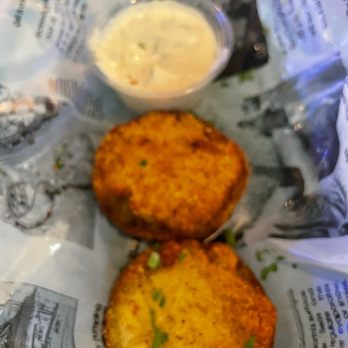 The Quarry Thornton - Fried green tomatoes.  They come with 6 slices. - Two fried green tomato slices with a golden-brown, crispy coating are positioned on paper, garnished with small green herbs. A small clear cup of creamy dipping sauce is placed above the tomatoes. The items are on a surface covered with printed paper, likely in a casual dining setting. - Bars Near Me - Thornton, Illinois