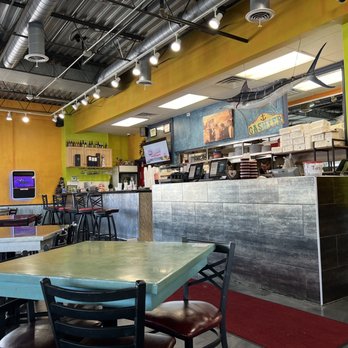 Las Islitas Stone Park - the interior of a restaurant - The image shows the interior of a seafood restaurant with vibrant yellow and green walls. A mounted fish decorates the wall behind the counter, which has a tiled front and kitchen staff working behind it. The ceiling has exposed ductwork and industrial lighting. There are tables and chairs for customers, with some tables visible in the foreground. A small TV and framed certificates are also present on the walls. - Mexican Near Me - Stone Park, Illinois