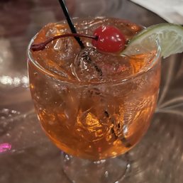 Singapore Sling - A cocktail with ice cubes is served in a clear glass, garnished with a red cherry and a lime wedge on the rim. The drink has an orange hue and is placed on a wooden table, with a blurred background that suggests a restaurant or bar setting.