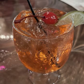 Trax - Singapore Sling - A cocktail with ice cubes is served in a clear glass, garnished with a red cherry and a lime wedge on the rim. The drink has an orange hue and is placed on a wooden table, with a blurred background that suggests a restaurant or bar setting. - New American Near Me - Scranton, Pennsylvania