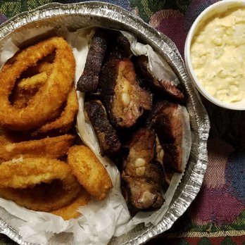 Mr E Chefs Barbecue - The image shows a round, aluminum takeout container filled with fried onion rings, grilled pork ribs, and a side of potato salad in a white cup. The onion rings are golden brown, and the ribs have a dark, charred exterior with visible bone and meat. The potato salad appears creamy with small chunks of potato. The container is placed on a multicolored, patterned fabric surface. - Caterers Near Me - Chicago, Illinois