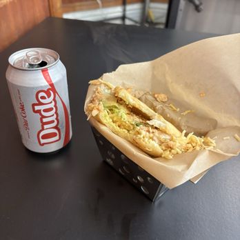 11 Degrees North - Chicken La Pelua Sandwich - arepa with fried plantains, Gouda cheese and garlic cilantro sauce - A can of Diet Coke and a sandwich in a paper-lined basket are positioned on a black table. The sandwich appears to contain lettuce, chicken, and cheese, with some shredded cheese on top. The background includes a brick wall, electrical outlets, and some metal furniture, indicating an indoor setting, possibly a casual dining or food preparation area. - Coffee & Tea Near Me - Chicago, Illinois