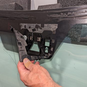 A Better Windshield - A person's hand, with a bandage on the finger, is holding a damaged windshield component inside a vehicle. The windshield has visible cracks and damage, and the dashboard and interior parts are partially visible. The image appears to be taken during a repair or inspection process. - Windshield Installation & Repair Near Me - Monument, Colorado