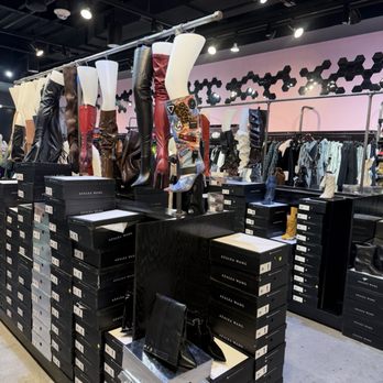Akira - The image features a retail store interior with mannequin legs displaying high-heeled boots in various colors and styles, placed on top of stacked shoe boxes labeled 'Azalea Wang.' The store has a modern design with a pink and black geometric wall pattern and clothing racks with hanging garments in the background. The ceiling is black with multiple spotlights illuminating the display. - Women's Clothing Near Me - Paramus, New Jersey