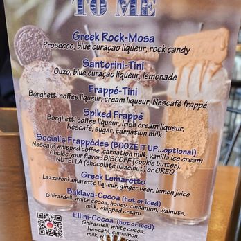 Caffe Social - The image shows a menu sign with a Greek theme, titled 'IT'S ALL GREEK TO ME.' It features various drinks and desserts such as Greek Rock-Mosa, Santorini-Tini, Frappé-Tini, Spiked Frappé, and social's Frappées, with detailed ingredient descriptions. The menu also offers hot or iced options for baklava-cocoa and ellini-cocoa, with a QR code at the bottom left corner. The sign is placed on a wooden table, with part of a metal holder visible at the bottom. - American Near Me - Norwalk, Connecticut