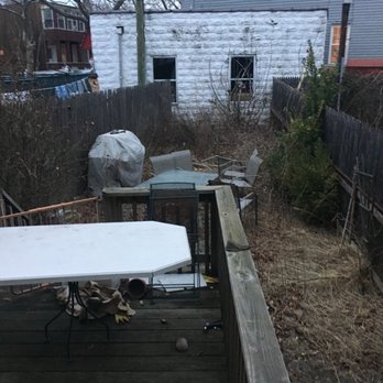 Smart General Contracting - Before - A backyard area features a wooden deck with outdoor furniture, including chairs and a table, alongside a covered grill. The yard is overgrown with dry grass and weeds, enclosed by a weathered wooden fence. In the background, there are neighboring houses, one with lit windows, and a small, unfinished white building with two windows and a door. The scene appears to be in late fall or winter, with leafless bushes and a generally unkempt appearance. - Stucco Services Near Me - Brooklyn, New York