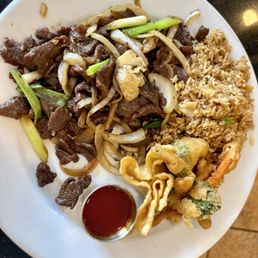 Bulgogi - The image shows a white round plate containing beef stir-fry with sliced onions and green onions, a portion of fried rice with small bits of vegetables, and assorted vegetable tempura including green vegetables and possibly shrimp, accompanied by a small cup of dark dipping sauce. The plate is placed on a dark surface, with a tiled floor visible in the background.