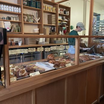 Two Seven Two - A bakery interior features a wooden counter with a glass display case containing pastries such as croissants, cinnamon rolls, and other baked goods. Behind the counter, a staff member wearing a cap and green shirt is working, with shelves of books, jars, and decorative items visible on the wall. The setting includes warm wooden accents and a partial view of the kitchen area. - Bakeries Near Me - Cincinnati, Ohio