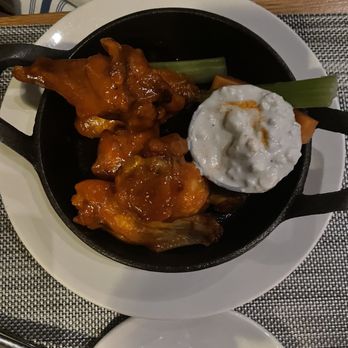 Half Moon - Buffalo Chicken Wings - The image shows buffalo chicken wings coated in sauce served in a black skillet placed on a white plate. Next to the wings is a dollop of creamy dip, possibly ranch or blue cheese, with a piece of green vegetable and a carrot stick. The setting appears to be a restaurant table with a textured placemat and a fork nearby. The background includes part of a person's arm and clothing. - New American Near Me - Dobbs Ferry, New York