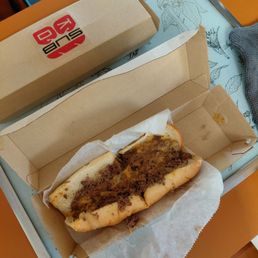 A partially eaten cheesesteak sandwich with beef, melted cheese, and bread is placed inside an open cardboard box on a tray. The tray has a decorative design and is on a wooden table. A sealed box with a red and black logo is also on the tray, and a gray cloth is partially visible in the corner.