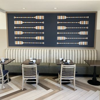 Anchor & Port - The image shows a dining space with a long, striped upholstered bench against the wall, three small square tables, and four chairs with woven backs. Each table has cups, saucers, and utensils. The wall behind the bench has a decorative art piece made of multiple paddle-shaped cutouts with colorful striped patterns. The flooring is a light-colored wood with a geometric pattern, and the ceiling has recessed lighting. The space appears clean and modern, suitable for casual dining. - Breakfast & Brunch Near Me - Oshkosh, Wisconsin