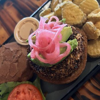 Twisted Root Burger - A burger with pickled onions and avocado on a bun, accompanied by potato chips, a slice of tomato, and a small container of dipping sauce. - Burgers Near Me - Bedford, Texas