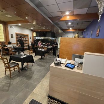 Cairo Kebab - Entrance - The image shows the interior of a restaurant with a mix of wooden and tiled flooring, and a ceiling with a combination of tiles and wooden panels. Multiple tables with black tablecloths are set for dining, with diners seated and being served by staff. The background includes a kitchen area with visible equipment and staff. Decor includes wall art and hanging light fixtures, creating a cozy dining atmosphere. - Middle Eastern Near Me - Chicago, Illinois