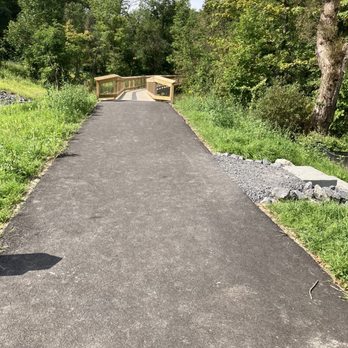 Carpenter Falls - Entrance to boardwalk to falls - A paved asphalt pathway leads to a small wooden bridge with railings, situated in a lush, green wooded area. The scene includes trees with dense foliage on both sides of the trail, and the sky is partly visible with some clouds. The pathway appears to be part of a park or nature reserve, providing access through the natural landscape. - Landmarks & Historical Buildings Near Me - Moravia, New York
