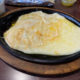 The image shows a cast iron skillet filled with melted cheese and cooked onions, placed on a wooden serving board. In the background, there are tortillas wrapped in foil and a small container of green sauce, indicating a Mexican or Tex-Mex dish. The setting appears to be a casual restaurant or taqueria.