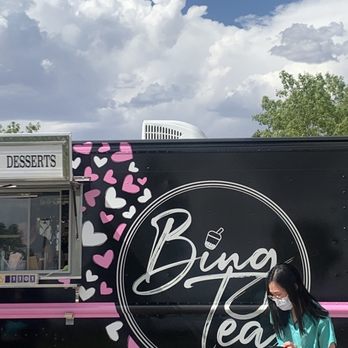 Bing Tea - A woman in a teal outfit and face mask stands beside a black dessert truck decorated with pink and white hearts, with a sign indicating desserts. - Desserts Near Me - Albuquerque, New Mexico