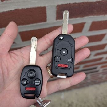 Eagle Locksmith - Car keys cut and programmed - A person is holding two car keys with remote controls in front of a brick wall. The keys have black plastic fobs with buttons for locking, unlocking, trunk release, and panic. One key has a keychain attached. The person is wearing a ring on their finger. The background features a red brick wall with gray mortar. - Keys & Locksmiths Near Me