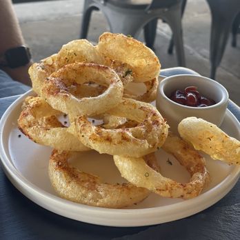 3128 Social House - Onion rings - A white plate holds a serving of golden, crispy fried onion rings, accompanied by a small cup of ketchup. The dish is placed on a dark table, with outdoor seating visible in the background, including metal chairs and a railing. The setting appears to be a casual dining area, possibly at a restaurant or cafe. - Gastropubs Near Me - Albuquerque, New Mexico