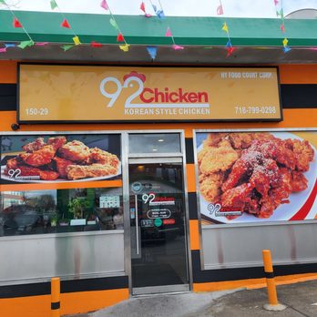 92Chicken - Store front facing Han Yang - The image shows the storefront of 92 Chicken, a Korean-style fried chicken restaurant located in Flushing, NY. The facade includes a large sign with the restaurant's name, contact number, and description, along with two large images of fried chicken dishes. The storefront has a black and orange color scheme, with a glass door and window. There are small orange bollards in front of the entrance, and a stop sign is visible on the left side of the image. - Korean Near Me - Flushing, New York