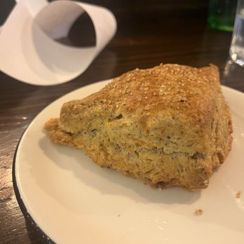 Ursula - Blue corn almond scone - A triangular-shaped blue corn almond scone with a golden-brown crust and a slightly coarse texture, topped with coarse salt. It is served on a white plate on a dark wooden table, with a glass of water and a green bottle in the background. The setting appears to be a restaurant or cafe environment. - New Mexican Cuisine Near Me - Brooklyn, New York