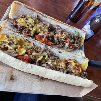 Craft'd - $29 2 foot Philly with about 1 foot of meat - Two Philly cheesesteaks with beef, peppers, onions, and melted cheese served on hoagie rolls, placed on a tray at a restaurant table. - American Near Me - Plainfield, Illinois