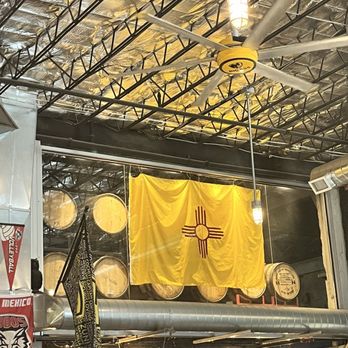 Rio Bravo Brewing - A yellow flag with a red Zia symbol is displayed inside a brewery, with barrels and industrial ceiling elements visible. - American Near Me - Albuquerque, New Mexico