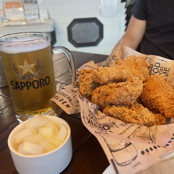 bb.q Chicken - Fried chicken pieces served on paper with a Sapporo beer and a bowl of pickled radish on a dark wooden table. - Korean Near Me - Markham, Ontario