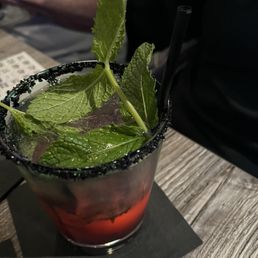 A cocktail featuring fresh mint leaves as garnish is shown in a glass with a salted rim. The drink is placed on a dark coaster on a wooden table, with a dimly lit background including a person in a black shirt and another person in a gray shirt. The setting appears to be a bar or restaurant.