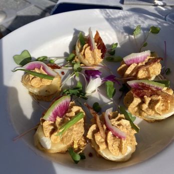 Devil's Kitchen - The best deviled eggs - A white plate holds several deviled eggs garnished with microgreens and thin slices of radish. The eggs are filled with a seasoned yolk mixture and have small pieces of bacon on top. The dish is set on an outdoor table with sunlight casting shadows, and a menu and utensils are visible in the background. - New American Near Me - Grand Junction, Colorado