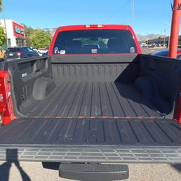 Look how beautiful this is - The image depicts the open cargo area of a red pickup truck parked in a parking lot. The truck bed is empty, with a textured black surface and sidewalls. The tailgate is lowered, revealing the truck's rear window and part of the cab. The background includes other parked cars, a building with signage, trees, and a clear blue sky. The scene is well-lit, indicating daytime.
