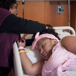 The Tidal Doula - Image: A support person holding the hair back of a birthing person laying in a hospital bed. The birthing person is wearing a pink gown.