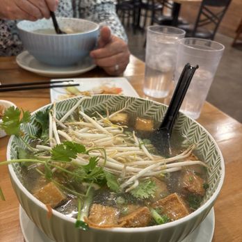 Wrap N Roll - Tofu glass noodle - A large bowl of Vietnamese noodle soup contains bean sprouts, tofu, and fresh herbs, served on a wooden table. Two glasses of water are nearby, and a person in a patterned shirt is eating from a smaller bowl in the background. The setting appears to be a casual restaurant with wooden furniture and a busy atmosphere. - Vietnamese Near Me - Albuquerque, New Mexico