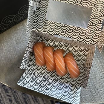 Fujisushi - Salmon nigiri - The image shows a decorative box with a black and white wave pattern, containing five pieces of salmon nigiri sushi. The sushi pieces are arranged in a row, each with a slice of salmon on top of rice. The box is open, revealing the sushi inside, and is placed on a wooden surface. The photo is taken from above, with part of a person's leg and shoe visible at the bottom of the image. - Sushi Bars Near Me - Eagan, Minnesota