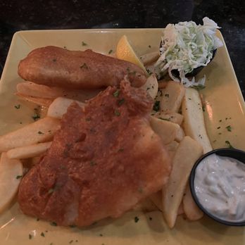 McCarthy's Pub - Fish and chips - A yellow square plate holds a serving of fish and chips, including a piece of fried fish, French fries, a lemon wedge, coleslaw, and a small container of tartar sauce. The dish is presented on a dark, possibly granite, table surface. - Irish Pub Near Me - Tequesta, Florida