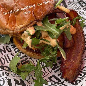 Piazza Messina - Love this place! It's a must try! Super cute patio, quaint atmosphere, & the food was delicious! - A breakfast sandwich with crispy bacon, fresh arugula, and a creamy sauce in a toasted bun, placed on branded paper at Piazza Messina. The sandwich appears freshly prepared with a variety of ingredients, including a green garnish and a toothpick for presentation. - Venues & Event Spaces Near Me - Cottleville, Missouri