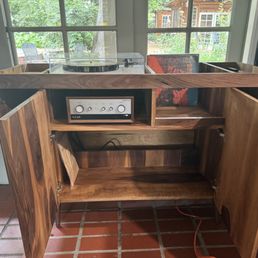 A wooden cabinet with two open doors reveals an audio amplifier inside, placed in a room with large windows that overlook a backyard with outdoor furniture and greenery. The cabinet is made of natural wood with visible grain patterns, and the interior contains a shelf and space for additional equipment. The room has a tiled floor and shelves with books and photographs on the right side.